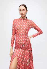 Robe ajustée à manches longues en tissu rouge transparent avec un motif de cœurs, présentant des détails froncés à la taille et une fente haute jusqu'à la cuisse.