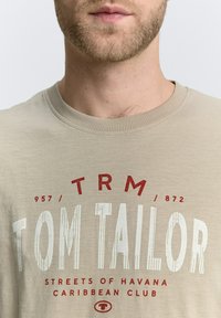 Beige katoenen t-shirt met ronde halslijn, voorzien van rode en witte tekst: "TOM TAILOR," "STREETS OF HAVANA," en cijfers. Gladde textuur.