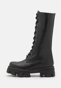 Stivale da combattimento in pelle nera alta fino al ginocchio con design di allacciatura frontale, punta rotonda e suola spessa e testurizzata con scanalature profonde.