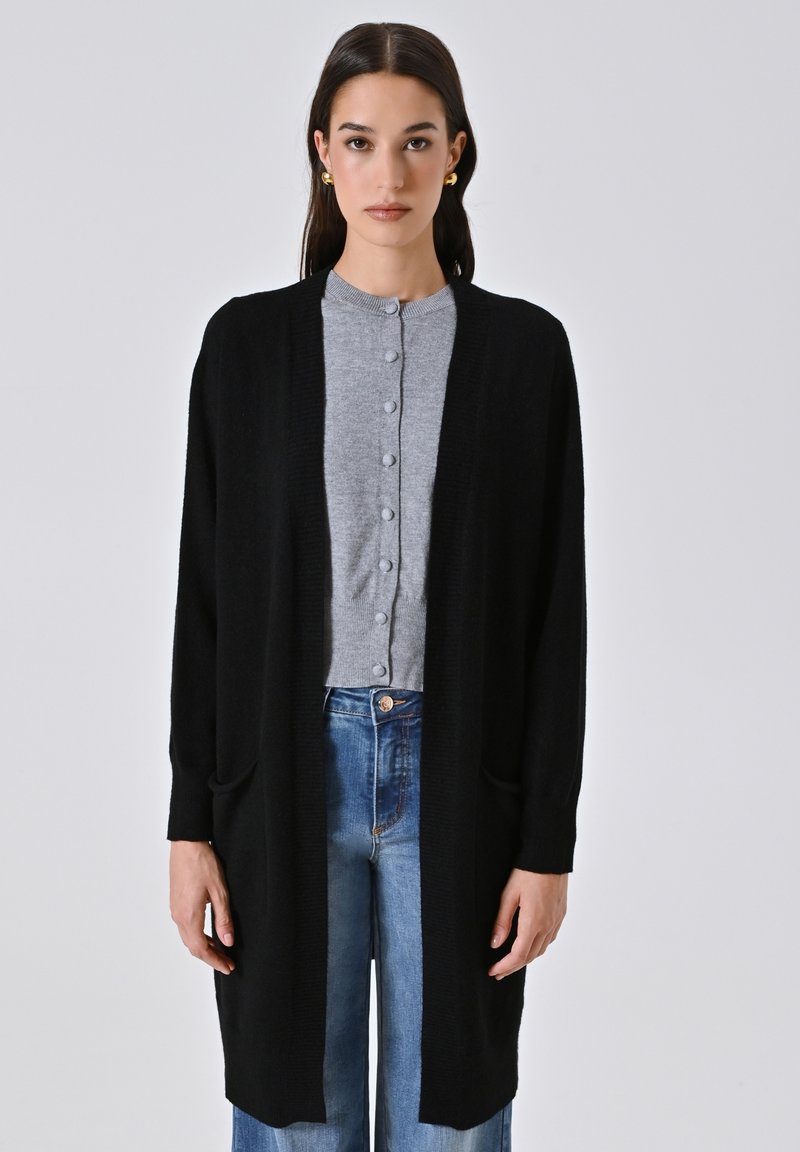 Frau, die einen langen schwarzen Cardigan über einem grauen, geknöpften Pullover und blauen Jeans trägt, steht vor einem schlichten hellen Hintergrund.