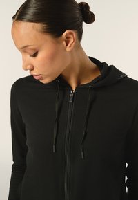 Felpa nera con zip, realizzata in un tessuto morbido, con cappuccio regolabile tramite lacci e zip metallica. Il design presenta accenti minimali vicino al collo.