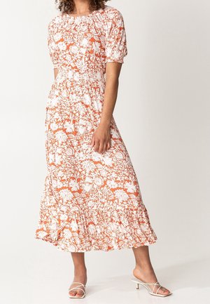 Kobieta nosząca białą i pomarańczową kwiecistą sukienkę midi z bufkami i falbanką u dołu, w połączeniu z białymi sandałami na obcasie z paskami.