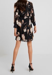 Robe noire à motifs floraux avec de longues manches bouffantes présentant des motifs floraux pastel. Tissu transparent, taille cintrée, longueur au-dessus du genou, portée avec des talons noirs.