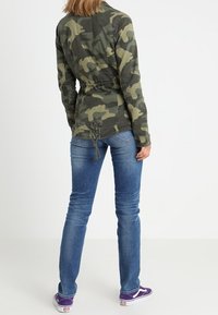 Maskáčová bunda v zelených tónech s elastickým pasem, dlouhými rukávy a texturovaným materiálem, kombinovaná s modrými džíny a fialovými teniskami.