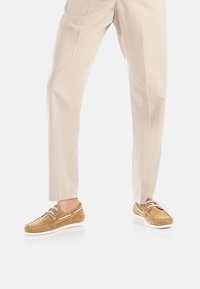 Scarpe da barca in suede marrone chiaro con suole in gomma bianca, dotate di occhielli e lacci argentati, indossate con pantaloni beige chiaro.