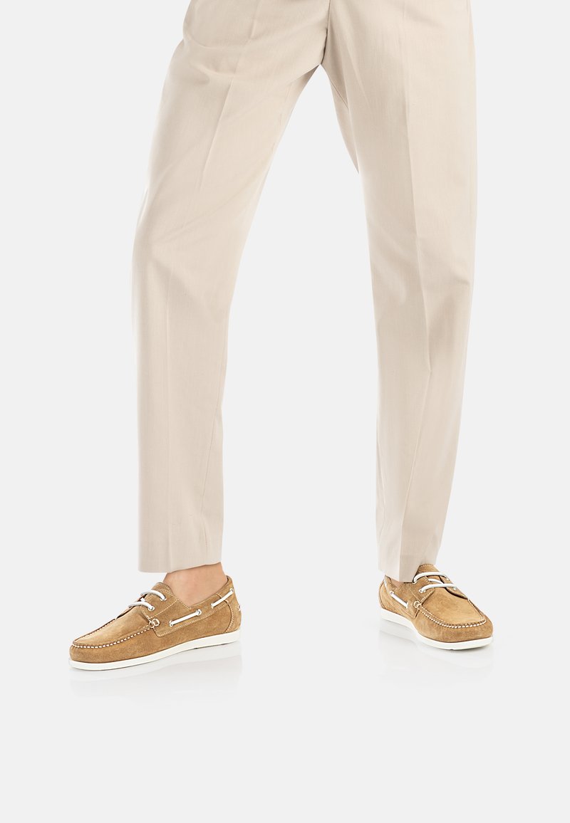 Scarpe da barca in suede marrone chiaro con suole in gomma bianca, dotate di occhielli e lacci argentati, indossate con pantaloni beige chiaro.