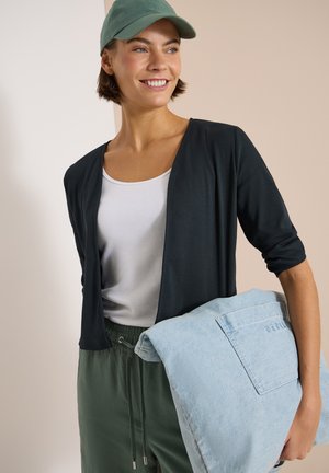 Kvinde smilende, iført grøn kasket, sort cardigan, hvid top og grønne bukser, holder en foldet lyseblå denimjakke med synlig lomme.