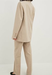 Beige skræddersyet jakkesæt med en struktureret blazer og lige bukser, der har en glat stoftekstur og knapper på blazerens ærmer.