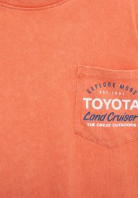 Tričko s krátkym rukávom v koralovej farbe, vyrobené z bavlny. Na prednej strane má náprsný vrecko s bielym a námorníckym potlačou: "TOYOTA Land Cruiser THE GREAT OUTDOORS".