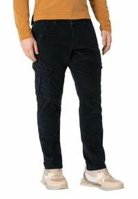 Dunkelbraune Corduroy-Cargohosen mit mehreren Seitentaschen, schmaler Schnitt und Knopfverschluss. Kombiniert mit hellen Sneakern, die einen strukturierten Stoff präsentieren.