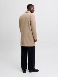 Beige uldblandingsfrakke med krave, rygslids, parret med sorte vide bukser og sort fodtøj. Glat tekstur, minimalistisk design.