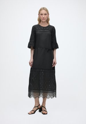 Heledate juustega naine, kes kannab musta midi-pikkusega kleiti pitsdetailide ja kellukese varrukatega, seisab valge ühevärvilise tausta ees mustade rihmikandadega.