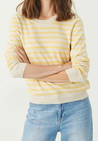 Femme les bras croisés portant un pull rayé beige et jaune et un jean bleu clair, sur un fond blanc uni.