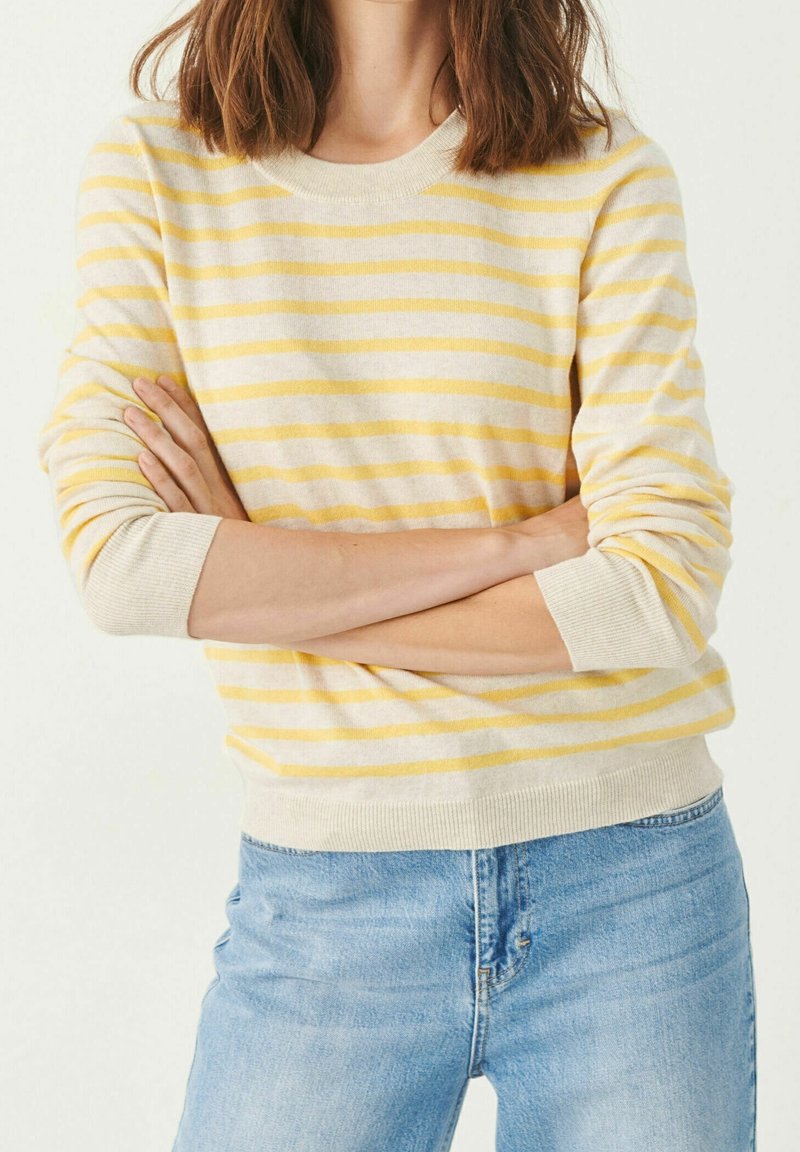 Femme les bras croisés portant un pull rayé beige et jaune et un jean bleu clair, sur un fond blanc uni.