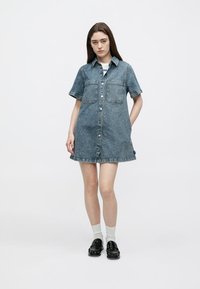 Φόρεμα πουκάμισο από τζιν σε ανοιχτό μπλε χρώμα, με κοντά μανίκια, δύο τσέπες στο στήθος και εμπρόσθιο κούμπωμα με κουμπιά. Το τελείωμα είναι ελαφρώς ξεφτισμένο.
