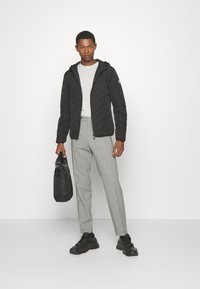 Svart quiltejakke med hette, lys grå skjorte, grå bukser og svarte joggesko. Modellen holder en svart veske. Enkelt design, nøytrale farger.