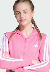 Sudadera rosa con cremallera y acentos de tres rayas blancas en las mangas, logo de Adidas. Tejido suave con un corte estándar y capucha.
