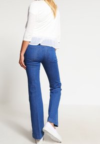 Lightweight white top with sheer bottom overlay, paired with blue denim flared jeans and white sneakers. Casual outfit from a rear view.