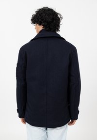 Granatowa wełniana kurtka z szerokim kołnierzem, zapinanymi mankietami i wyraźnie zbudowanymi ramionami. Gładka faktura, minimalne detale, dopasowany krój.