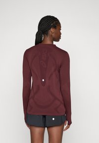 Camisa deportiva de manga larga color burdeos con diseño texturizado en la espalda y agujeros para los pulgares en las mangas. Combinada con pantalones cortos negros.