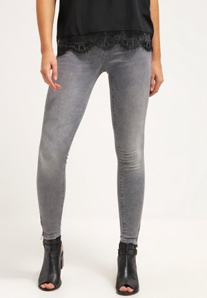 Femme portant un jean skinny gris avec des détails délavés, un haut noir orné de dentelle, et des bottines noires à bout ouvert.