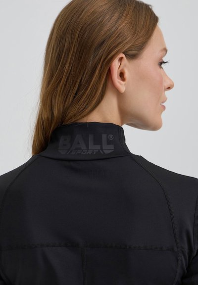 Moters šviesiai rudos spalvos plaukai, vilkinti juodą sportinį marškinėlį aukštu apykaklės apvadu, ant nugaros su "BALL SPORT" logotipu, žiūrinti į dešinę.