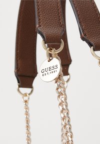 Hnědý kožený popruh na kabelku se zlatými kovovými prvky, s dekorativním řetízkem a značkovou kovovou visačkou s nápisem "GUESS EST.1981."