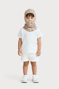 Kisgyermek fehér pólót és rövidnadrágot visel, fehér sportcipővel, valamint egy barna mintás balaklavát, amely a fejét és nyakát takarja, egy egyszínű háttér előtt áll.