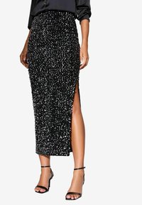 Sort midikjole med palietter, der har en tætsiddende pasform, gennemsigtig sideslids og glat tekstur, parret med sorte strappy sandaler.