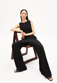 Schwarzer ärmelloser Jumpsuit mit tailliertem Oberteil und weitem Bein, kombiniert mit strukturierten schwarzen offenen Schuhen mit Absatz. Holzstuhl im Hintergrund.