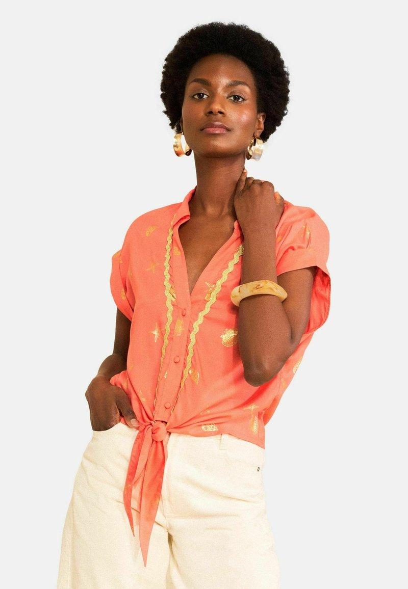 Femme aux cheveux courts naturels portant une chemise corail boutonnée nouée à la taille avec des motifs dorés, un pantalon ivoire, des boucles d'oreilles créoles dorées, et un bracelet marbré.