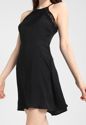 Robe noire à bretelles avec des accents de dentelle au niveau du décolleté, un bustier ajusté et une jupe évasée avec un ourlet arrière légèrement plus long.