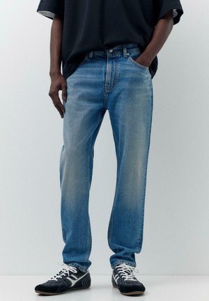 Osoba oblečená v vyblednutých modrých džínsach, čiernom tričku a čierno-bielych šnurovacích teniskách, stojaca na jednoduchom svetlom pozadí.