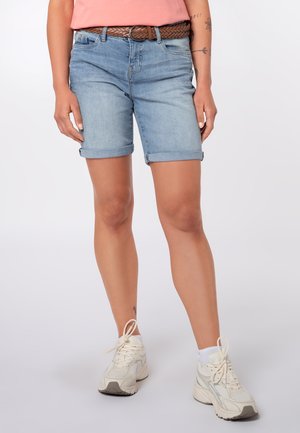 Person trägt hellblaue Jeans-Shorts mit umgeschlagenem Saum, einen geflochtenen braunen Gürtel, weiße Sneakers und ein pinkes Oberteil, steht vor einem einfarbigen Hintergrund.