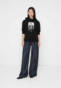 Fekete kapucnis póló, rajta egy öltönyös személy grafikájával. Széles szárú sötét farmernadrággal és fekete cipővel párosítva. Simább anyagtextúra.