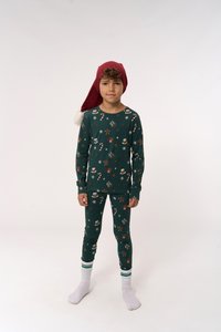 Grünes Pyjama-Set mit festlichen Mustern von Lebkuchenmännern, Schneeflocken und Zuckerstangen. Kombiniert mit einem roten Weihnachtsmann-Hut und weißen Socken.
