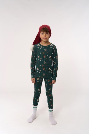 Grünes Pyjama-Set mit festlichen Mustern von Lebkuchenmännern, Schneeflocken und Zuckerstangen. Kombiniert mit einem roten Weihnachtsmann-Hut und weißen Socken.