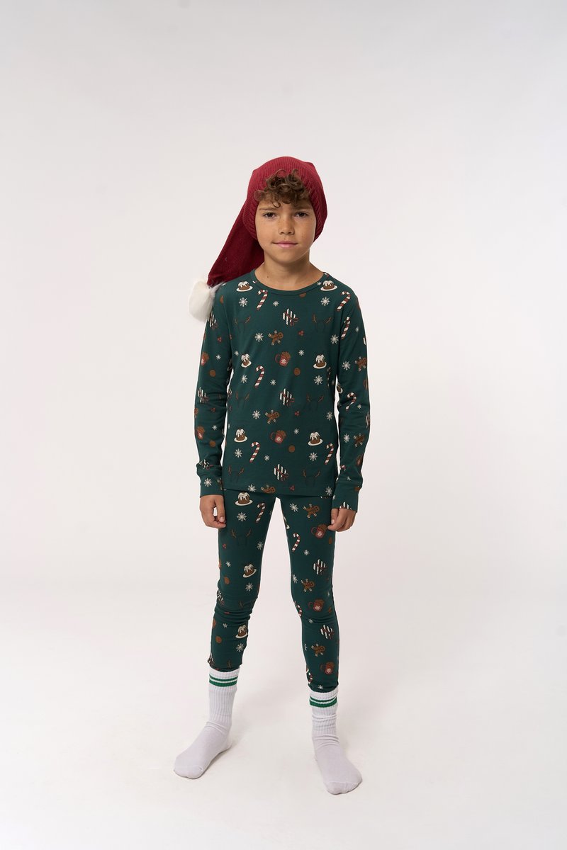 Grünes Pyjama-Set mit festlichen Mustern von Lebkuchenmännern, Schneeflocken und Zuckerstangen. Kombiniert mit einem roten Weihnachtsmann-Hut und weißen Socken.