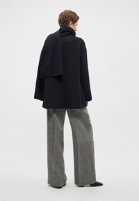 Nadměrný černý svetr s vysokým límcem, detailem skládajícího se šálu, doplněný širokými šedými kalhotami s kostkovaným vzorem. Hladká textura po celém povrchu.