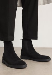 Svart mockaskinn Chelsea-boots med en texturerad finish, rund tå och platt gummisula; i kombination med skräddarsydda svarta byxor.