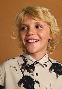 Chemise à manches courtes en tissu léger, avec une base beige ornée de motifs abstraits noirs, et un col classique avec fermeture par boutons.