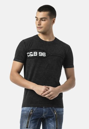 Jonge man in zwart t-shirt met witte "C&B 96" tekst en blauwe spijkerbroek met ritssluitingen aan de voorkant, kijkt naar links tegen een effen achtergrond.