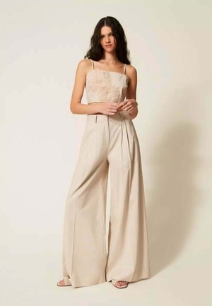 Giovane donna con capelli lunghi e scuri indossa pantaloni beige a gamba larga e un top beige senza maniche con texture floreale, in piedi contro uno sfondo chiaro.