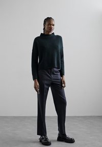 Maglione verde scuro, texture a trama, con collo alto, abbinato a pantaloni navy su misura e scarpe nere. Modello in piedi davanti a uno sfondo neutro.