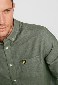 Groen button-up shirt gemaakt van textuurstof, met een borstzak met een gouden geborduurd logo en witte knopen.
