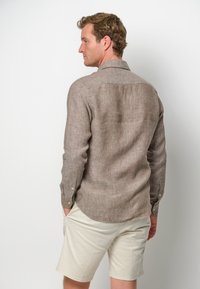 Camicia lunga in lino marrone con colletto, polsini con bottone e orlo curvo, abbinata a pantaloni corti di colore chiaro.