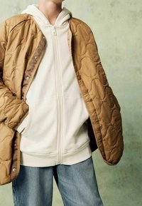 Personne portant un sweat à capuche crème zippé et un jean bleu clair avec une veste matelassée marron drapée sur les épaules.