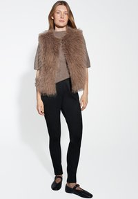 Gilet in faux fur marrone su un maglione beige a trama, abbinato a pantaloni neri aderenti e ballerine nere. Design semplice, materiali morbidi e colori neutri.