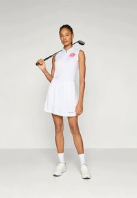 Λευκό γυναικείο γκολφ φόρεμα χωρίς μανίκια με γιακά, ροζ λουλουδάτο λογότυπο στο στήθος, φούστα που φουσκώνει και ταιριαστά λευκά αθλητικά παπούτσια. Κλαμπ γκολφ κρατημένο στον ώμο.