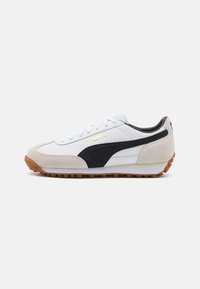 Zapatilla blanca con una franja de ante negro, detalles de ante gris y una suela de goma con un patrón texturizado. Logotipo dorado en el lateral.